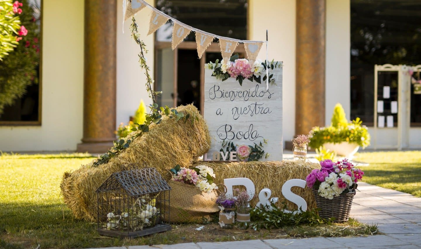 Entrada decorada con dos pacas de paja, unas iniciales P y S, flores, y un cartel en el que se puede leer "Bienvenidos a vuestra boda"