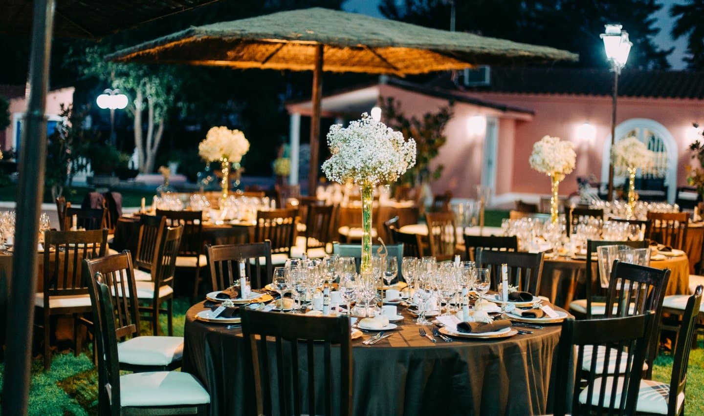 Mesa con 9 sillas, cubertería y vasos con una gran flor en el centro. Al fondo se pueden observar un parasol sobre unas mesas.