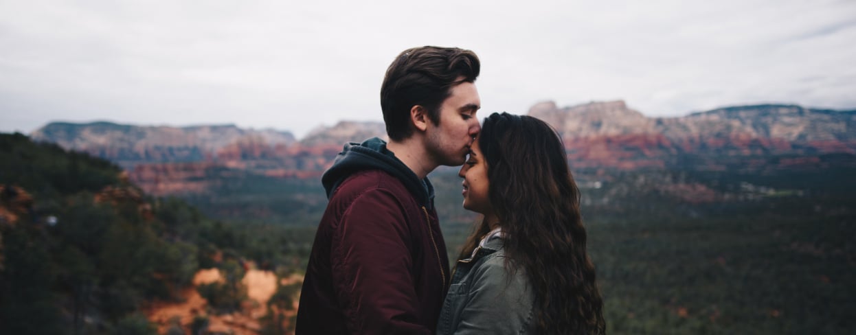 Imagen de una pareja en la montaña con vistas de fondo mientras el chico besa en la frente a la chica